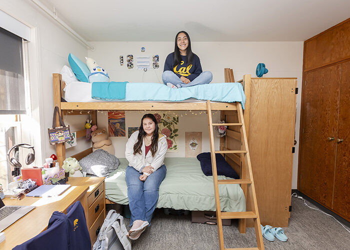 Two smiling students sit on their bunk bed; one student is on the top bunk and the other sits on the bottom bunk. The bed is positioned between a desk and dresser on the left, and a closet space on the right side.