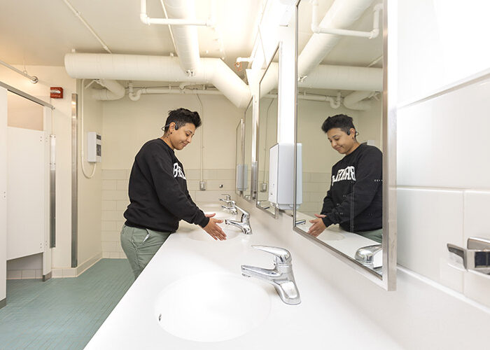 A student washes their hands at a sink in a shared bathroom that features individual stalls, sinks, and a mirror.