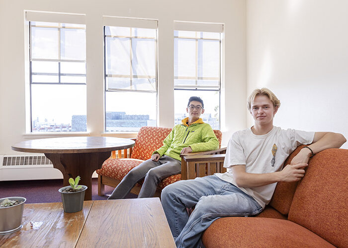 Interior shot of a shared living room featuring two students sitting on couches next to two small tables.