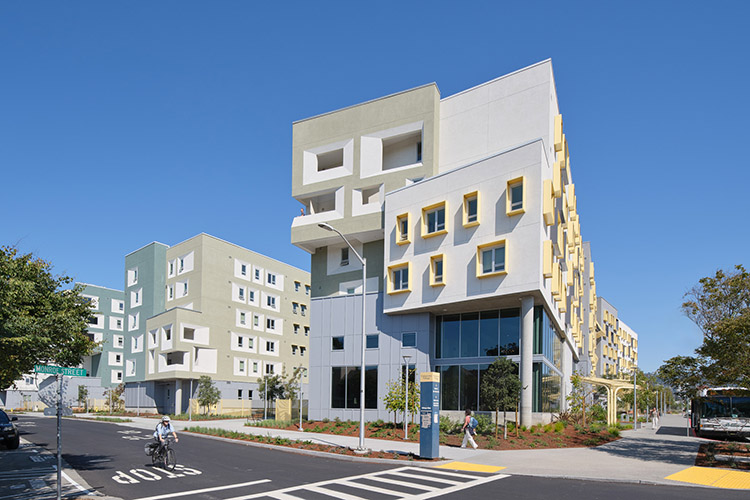 Exterior shot of a xučyun ruwway buildings on a sunny day. One person on a bicycle rides across the parking lot, and another walks on the sidewalk alongside the building.