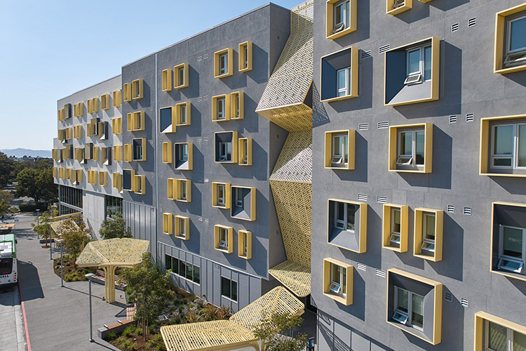 A xučyun ruwway apartment building with many windows is shown next to an outdoor patio. An AC Transit bus is parked outside next to the patio.