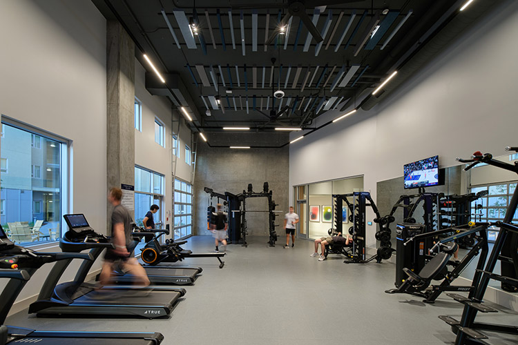 Several people are exercising in the fitness center. One person is walking on a treadmill, while the others are exercising near the weight machines on the other side of the room.