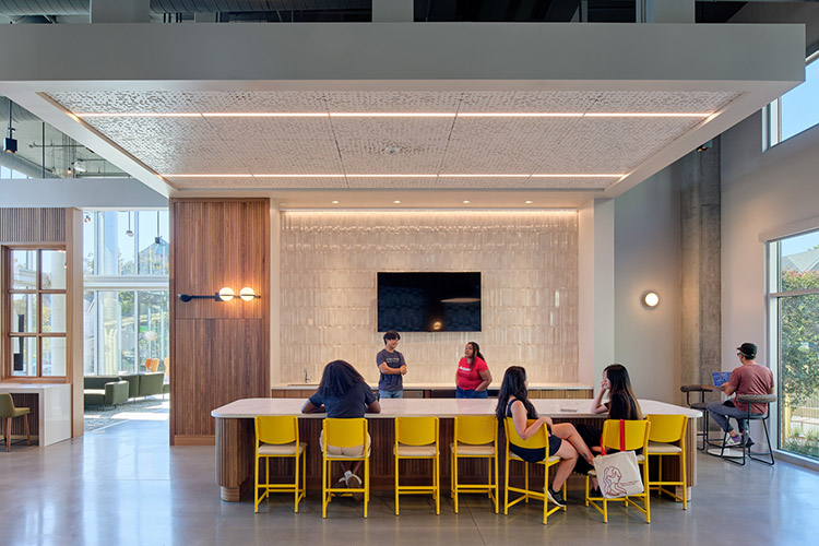 Several students sit at a long table in a communal kitchen. Two other students stand behind the table next to the kitchen counter. A TV is mounted on the wall.