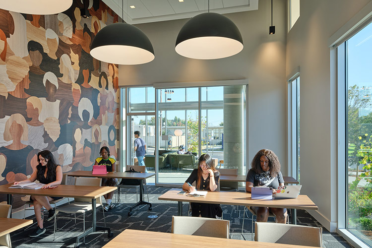 Several students sit at tables inside a communal study space. There are large windows and glass doors in the room.